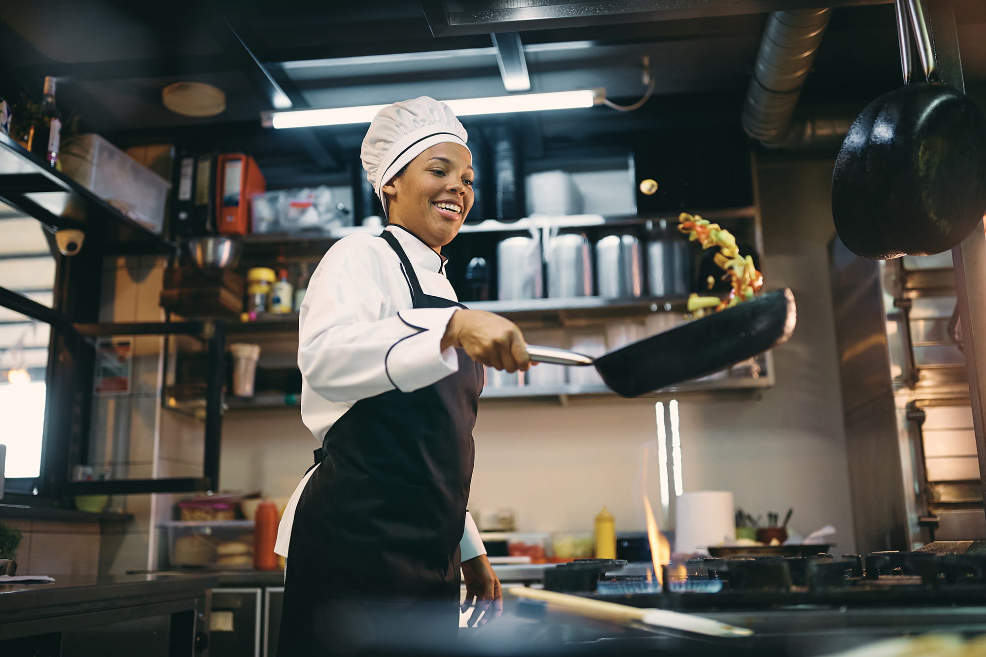 A smiling female chef in a restaurant kitchen sauteing ingredients as she flips them in the air.  