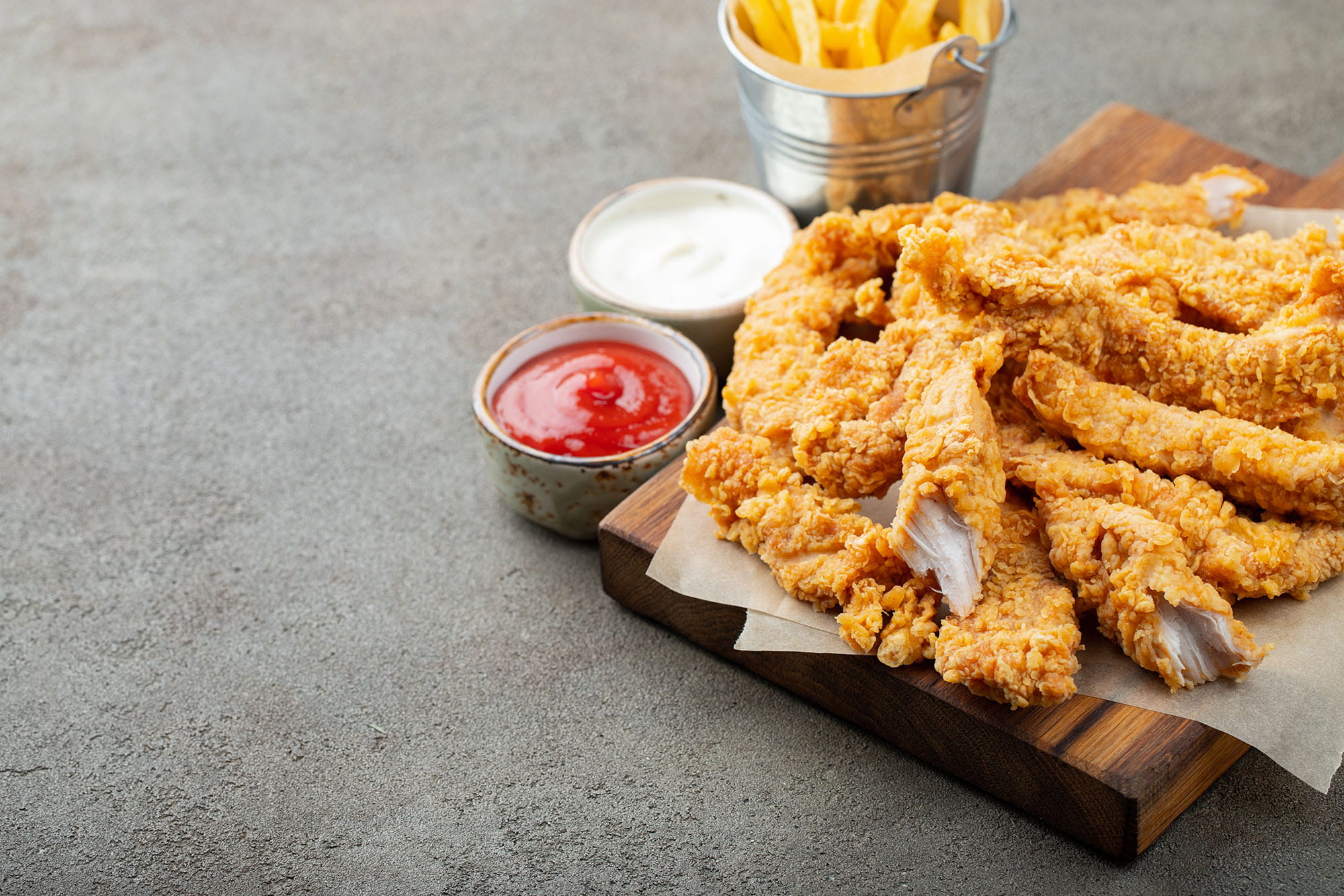 Wood cutting board with fried chicken tenders on top and sides of dipping sauce and French fries.