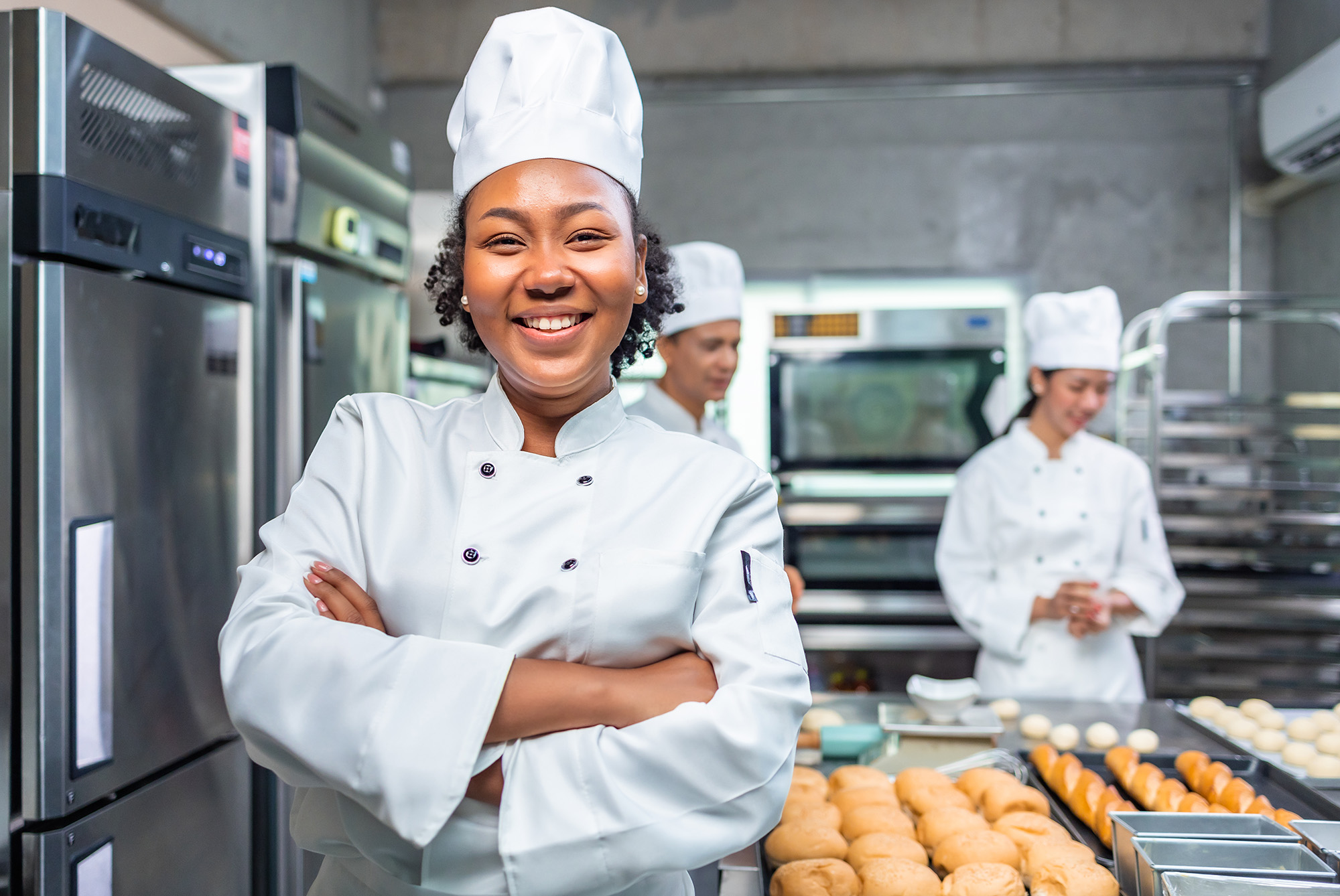  A smiling female baker faces the camera, with trays of baked goods that are in soft focus to her side. 